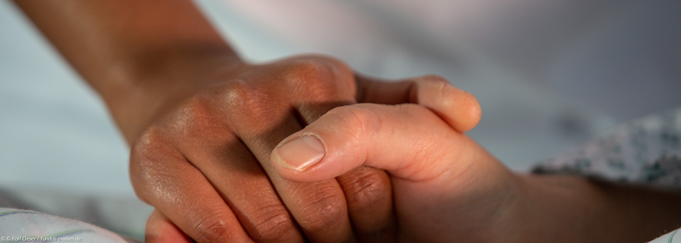 Eine Person liegt auf einem Bett. Ihre Hand wird von einer anderen Person gehalten. Auf dem Bild sind nur die Hände zu sehen.